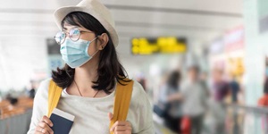 A woman with glasses wearing a hat and a mask, holding her passport and the straps of a yellow backpack.
