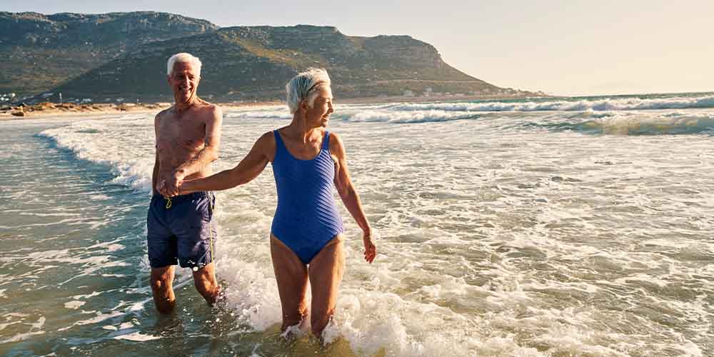 A senior man and woman walk along a beach holding hands while knee-deep in a body of water. There are small waves coming in with white caps and the water is greenish-blue. The man is wearing blue swim trunks, no shirt and has short white hair. The woman is wearing a purple one-piece bathing suit, has short whte hair and is looking toward the water. You see a mountain spanning the horizon. 