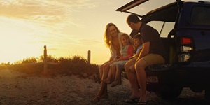 A woman, two small children and a man sit on the back hitch of a truck, with the trunk door raised above, on a sandy beach. The woman on the far left has long blonde hair and is wearing an orange shirt, beige sweater and is barefoot. She is looking toward a child on her right, who wears her blonde hair in a ponytail and is wearing a pink and green sweater with jean shorts and is also barefoot. Next to her is a child with short brown hair, and a man with short brown hair, wearing beige shorts and a brown T-shirt. The man is looking toward the children in the middle and his back is turned. The truck is blue with red and white tail lights and a yellow licence plate. In the background there is a sunset of yellow and orange and a wooden fence with some greenery.