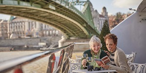 Two people sit at a white iron table by a body of water, looking at a red and white book. There is a small evergreen plant behind them. The one on the left has short, white hair and is wearing a green floral scarf and a dark green cardigan. They are holding a glass of white wine. The person on the right has short brown hair and is wearing a striped brown top and a pleated light brown sweater. They are both smiling and looking down at the book. There is a second glass of white wine on the table alongside an analog camera. In the background you see a blurry green iron bridge stretching over the body of water on the left as well as the building behind.  