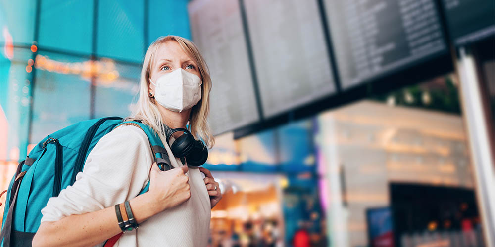 A woman with shoulder length blonde hair is staring into the distance. She is wearing a N95 mask. She has a pair of black earphones around her shoulders. She is wearing a teal backpack with grey straps, which she is holding onto with her hands. She appears to be standing in an airport. Behind her, overhead is a screen with a list of destinations and information that are blurred out.
