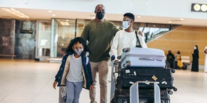 Three people, two adults and a young school-aged girl, each wearing masks are walking. A man, in an olive green sweater is rolling a grey suitcase behind the little girl. A woman wearing glasses is pushing a cart filled with three suitcases stacked on top of each other.