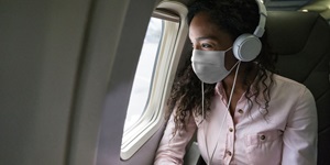 A woman with long curly hair, wearing a light pink button up shirt is wearing a white mask and white headphones. She is seated on an airplane, looking out the window. 