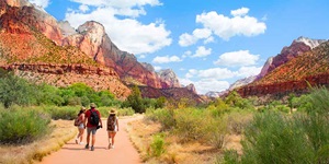 Three people walk with backpacks, shorts and hats along a dirt road in the desert surrounded by green vegetation and dried grass. There are mountainous cliffs of varying shades of red, white and brown on either side of the path stretching far into the distance. The sky is blue with white clouds.