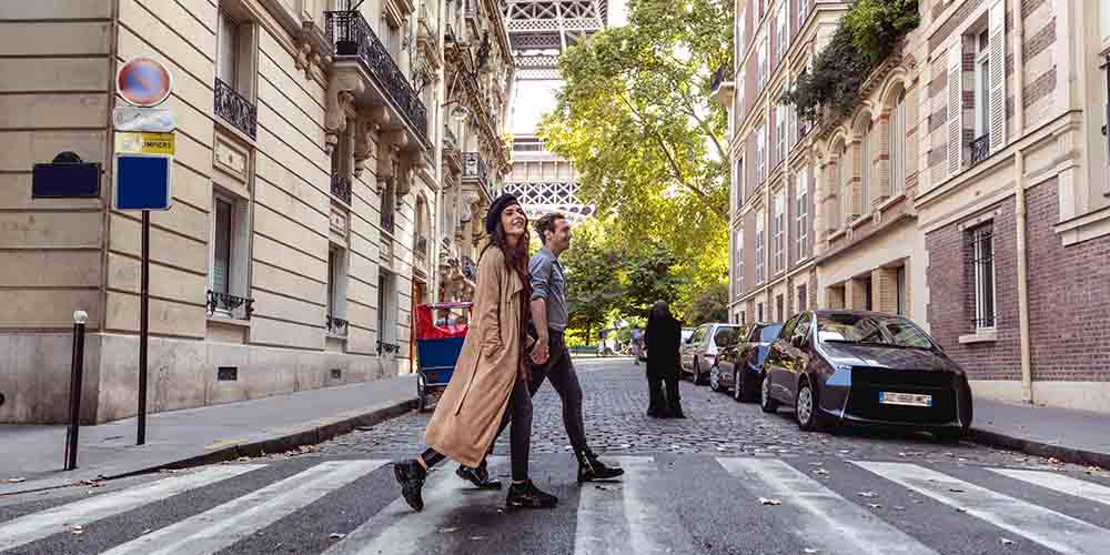 Two people walk across a crosswalk with painted horizontal white lines on the road. The one closest to the camera is wearing a long brown trench coat, black pants, black shoes and a black beret hat, and has long brown hair. Beside them is a person with a grey sweatshirt, black pants and black boots and has brown hair in a bun. They are holding hands. A line of cars is seen on the right side of the street behind them and historical architecture buildings surround. A glimpse of the Eiffel Tower is shown in the distance among green foliage.  