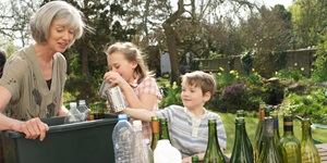 A woman holds a recycling bin while two young children toss in recyclables, plastic and glass bottles stacked in front of them