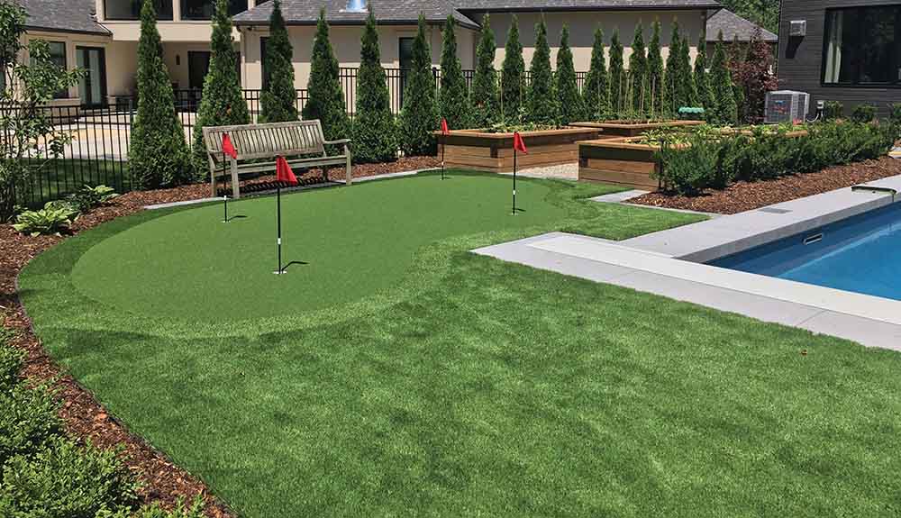 A backyard golf course made with artificial turf is seen next to a pool, a row of small, neat trees behind the golf course