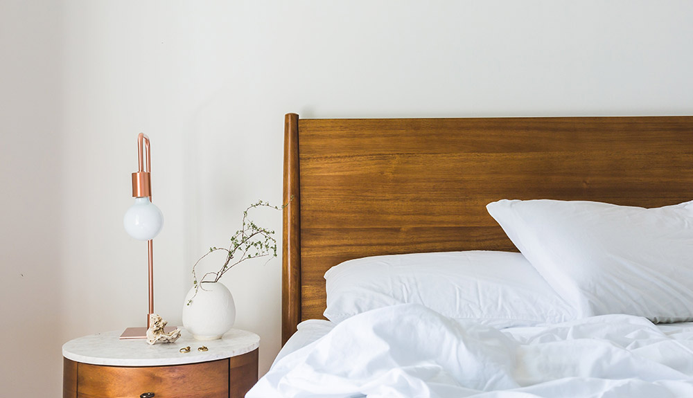 Night stand with a lamp and plant next to an unmade bed