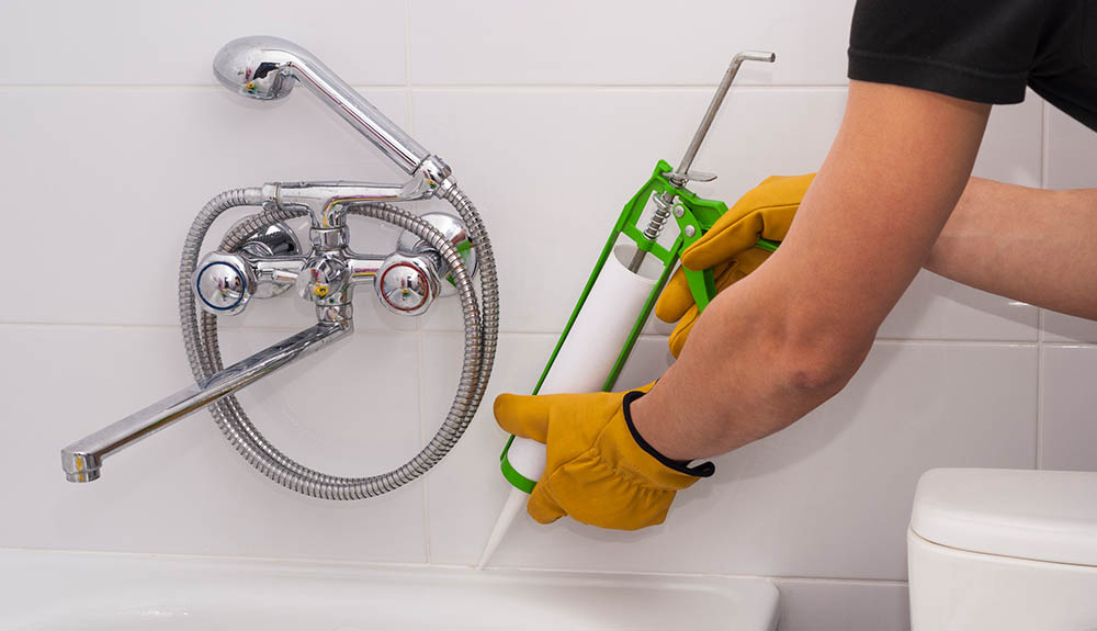 Man works caulking gun at the top of his tub beneath the faucet