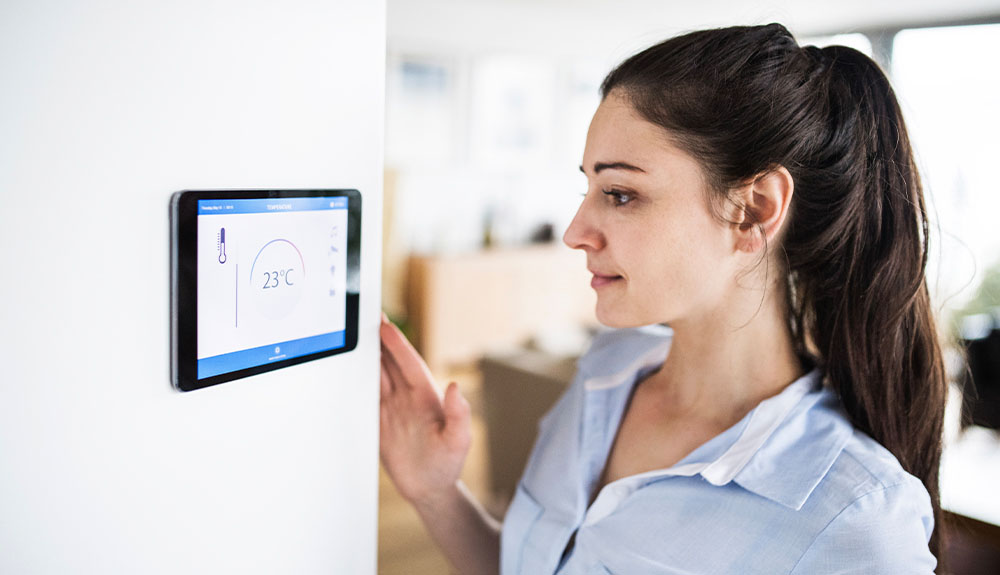 A woman stands in front of a smart thermostat mounted on a wall.