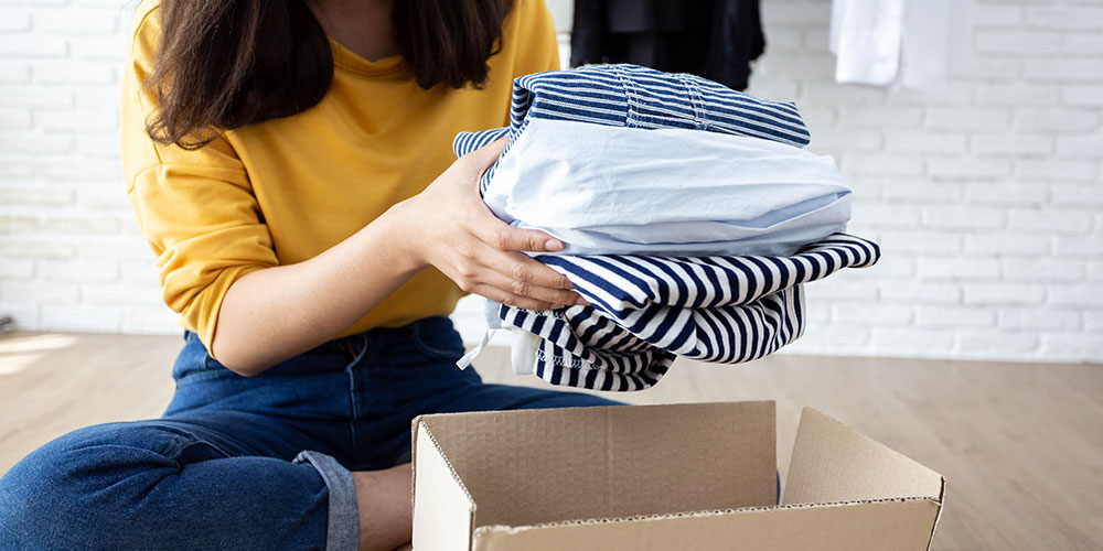 Woman in yellow top and jeans sits on the floor as she puts a stack of clothes into a box