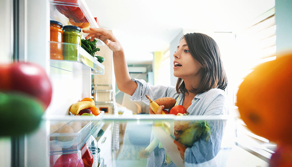 Woman with an armful of produce reaching at the door of her opened fridge