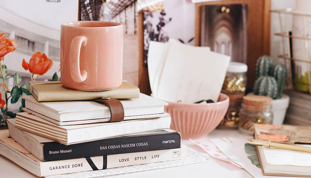 Stack of books and notebooks with a pink mug, resting on a busy desk