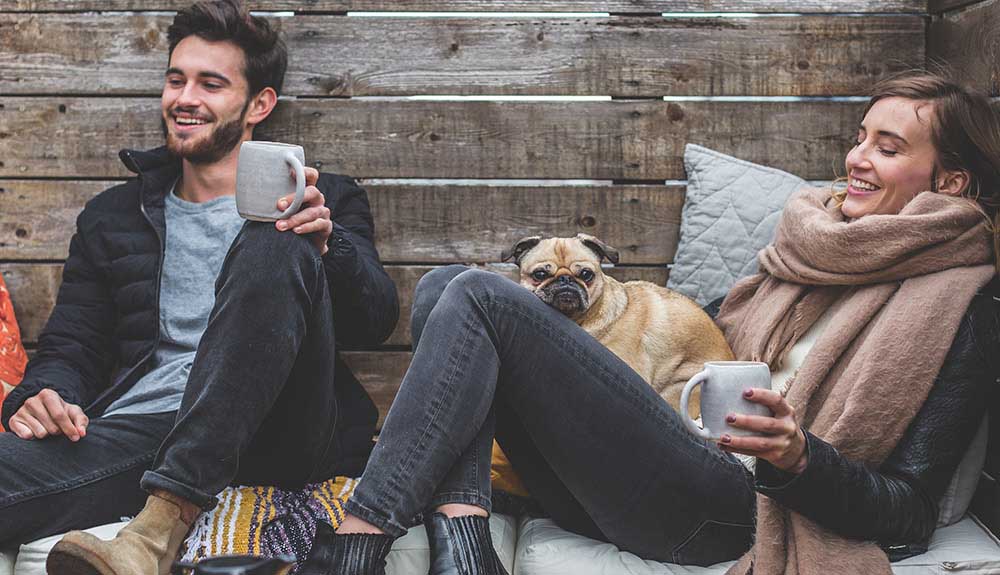 Man sits with a mug on his knee as a woman sits holding a mug with a pug on her lap outside on a cozy patio