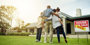 Family of four standing on lawn in front of house with Sold sign