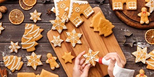 Hands piping icing onto snowflake cookie over background of wooden surface scattered with other festive cookies