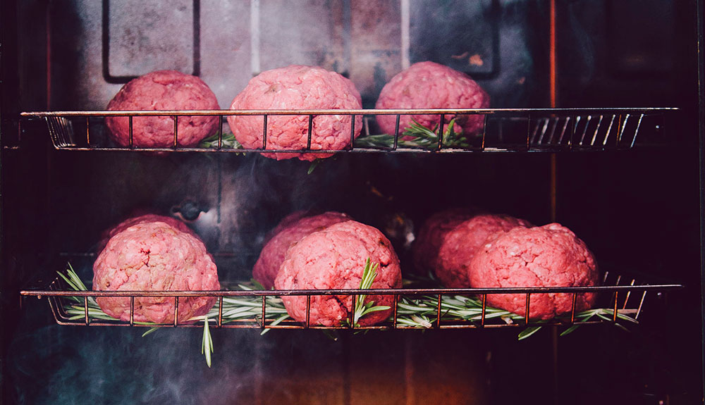 Two rows of large meatballs on herbs inside of a smoker