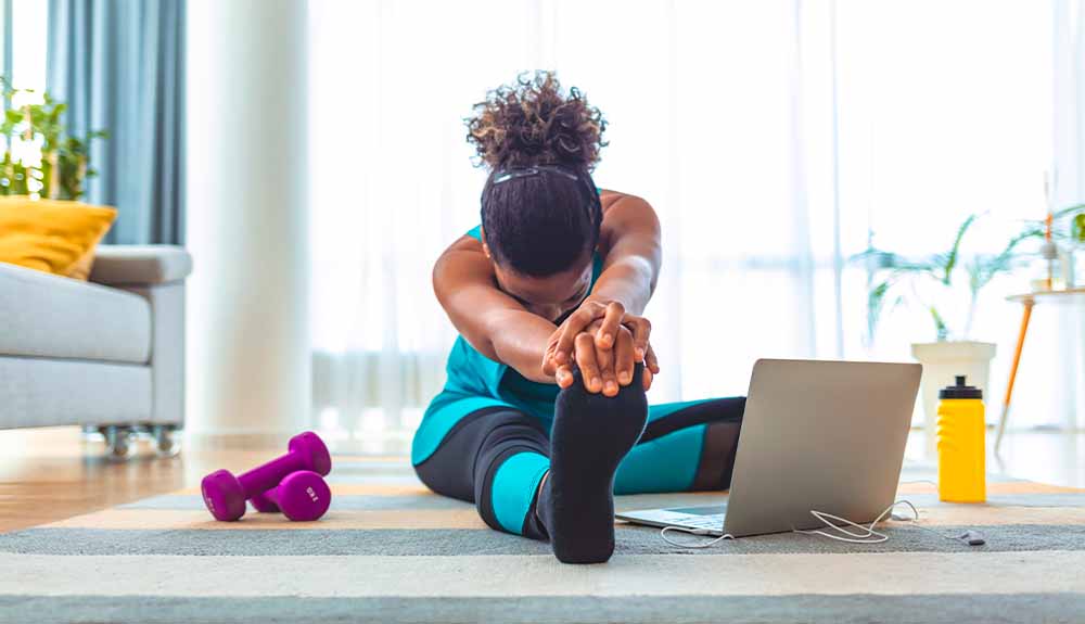 A woman sits on her living room floor stretching in front of a laptop