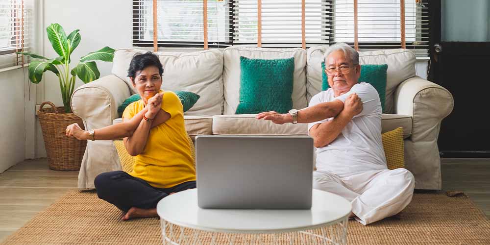 An older man with an old lady on a yoga mat looking at a laptop screen to stay in a fitness