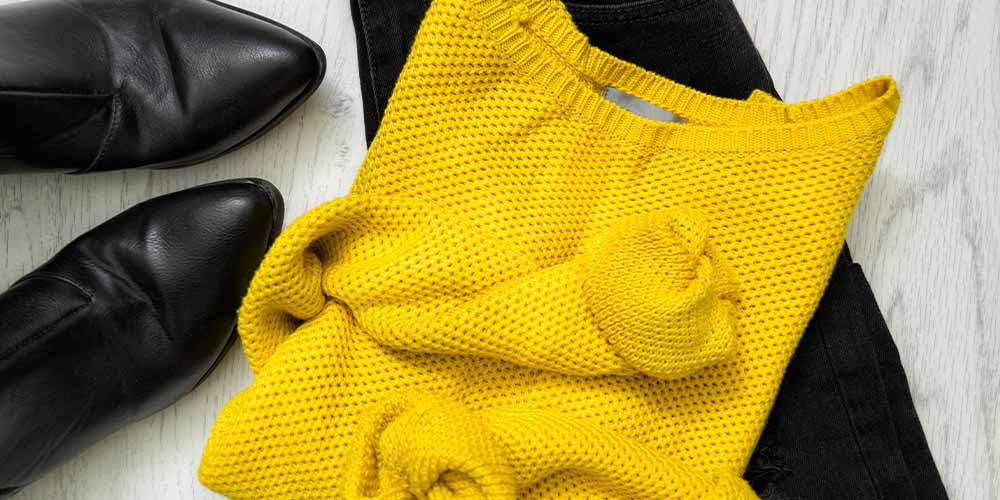 A mustard-coloured sweater is folded on top of black pants, next to a pair of black ankle boots
