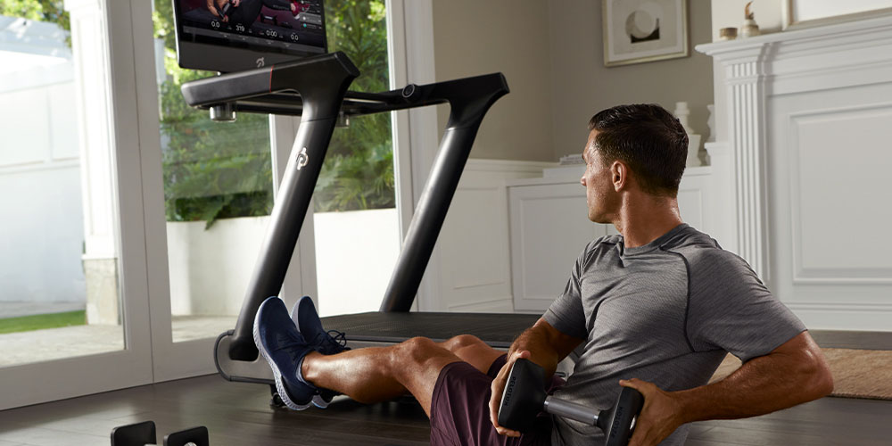A man does a mat work routine next to a Peloton Tread machine in his home