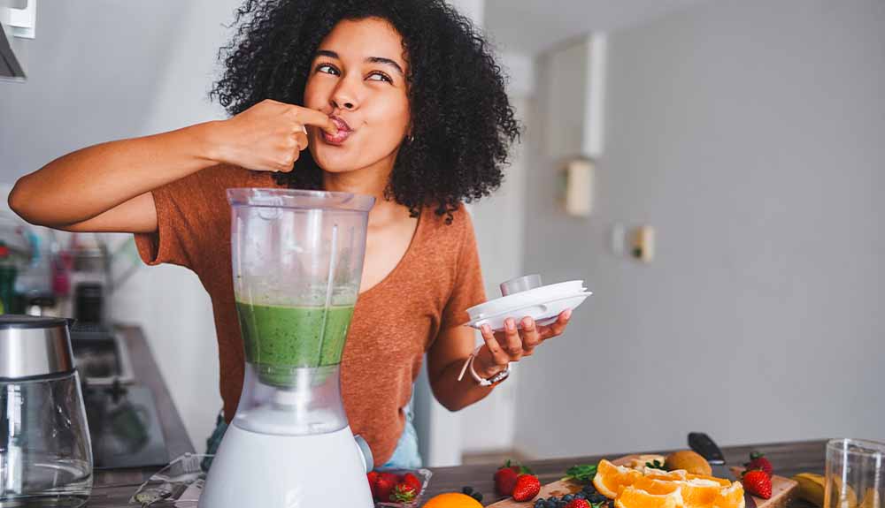 A woman is shown licking her finger, a blender with a green drink in it sits in front of her