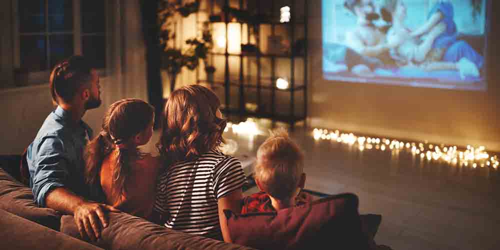 A family sits on a couch watching a movie projected onto their living room wall