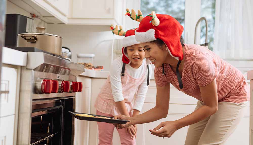 A mother and daughter with matching reindeer hats put a tray of cookies in the oven 