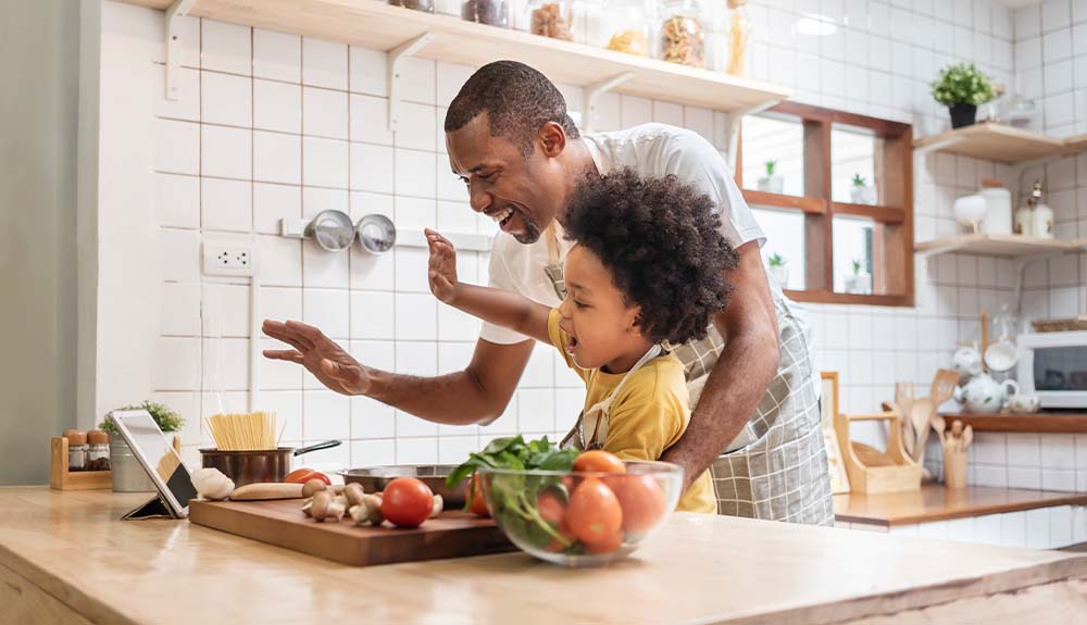 A father and daughter are shown in a kitchen with vegetables on the counter