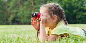 A young girl is shown lying in the grass looking through red binoculars