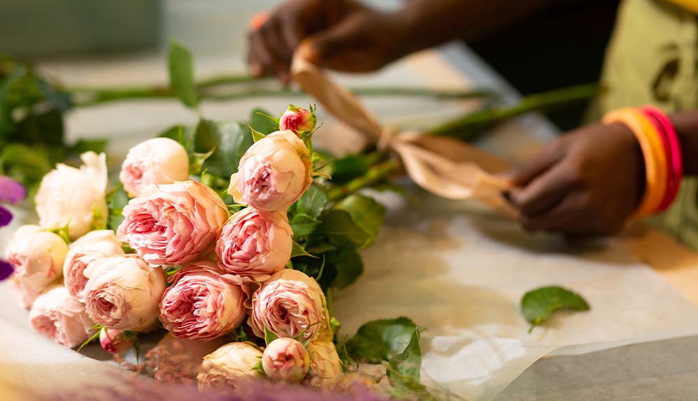 A bouquet of pale pink flowers lie flat on a table while a set of hands tie a bow around their stems