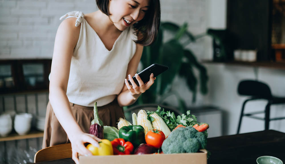 A woman looks at her phone as she unpacks a box of produce in her kitchen