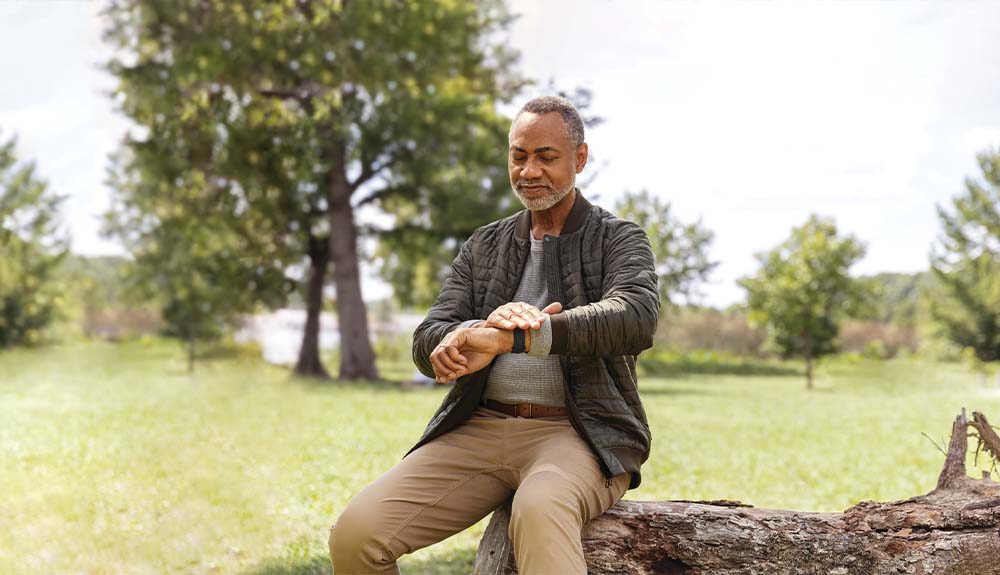 A man sits on a log in a rural setting as he looks at a fitness tracker on his wrist