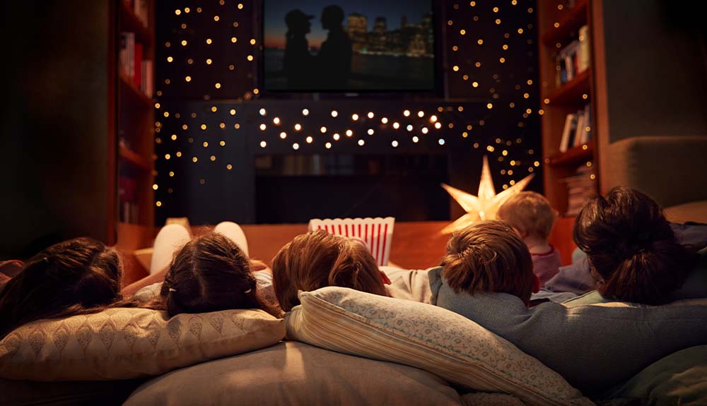 A family of five is shown from behind while they lie on the floor and watch a movie in their living room