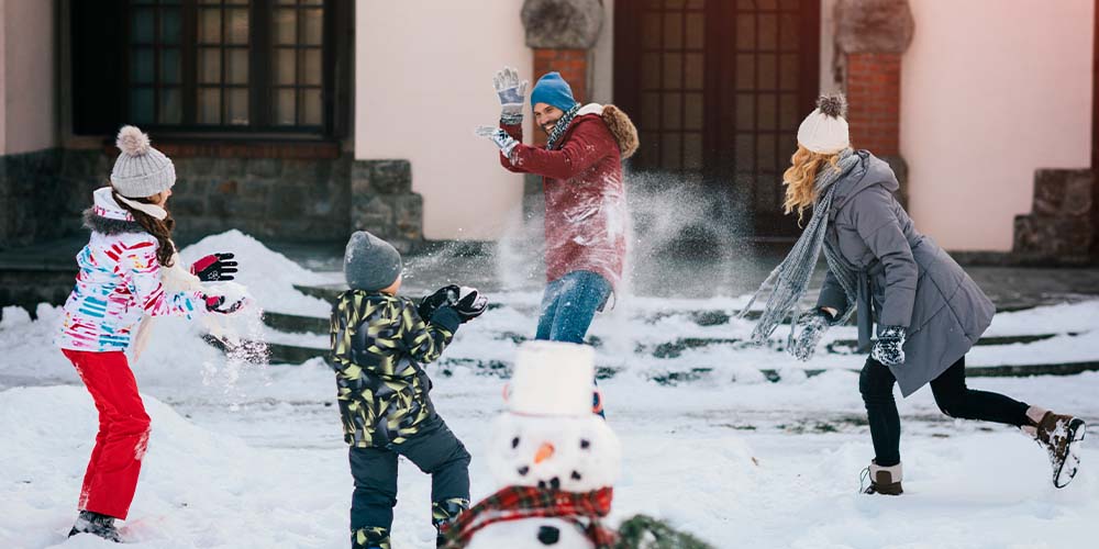A family has a snowball fight in front of their house with a snowman in the foreground