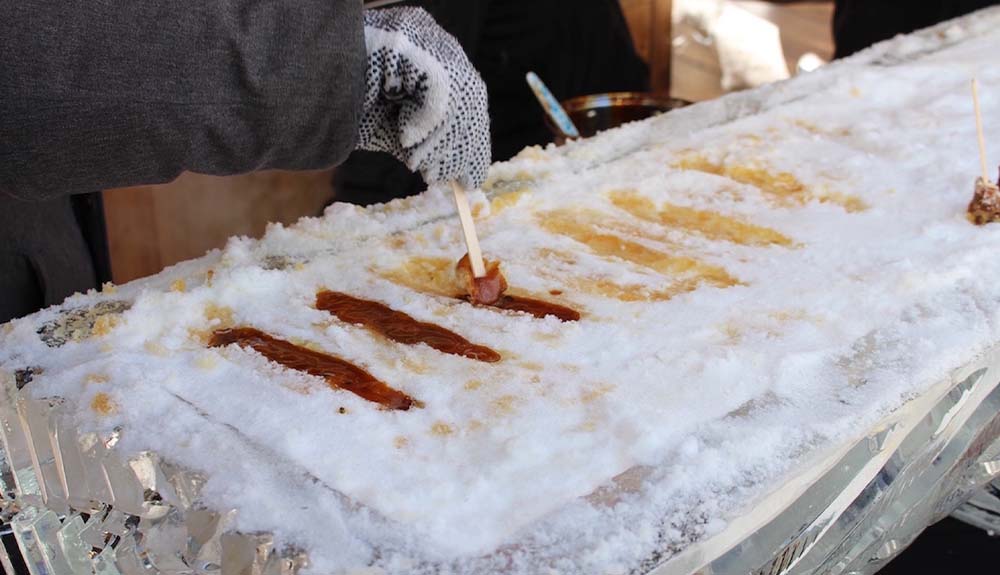 Strips of maple syrup are shown on a long tray covered in snow with a hand rolling one strip with a popsicle stick