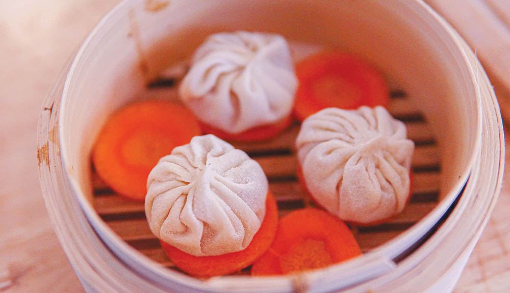 Dumplings are shown on top of carrot coins in a steamer basket