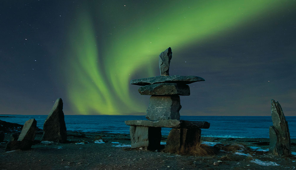 Green northern lights are shown in a swirl in the night sky with an inuksuk in the foreground