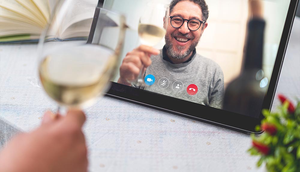 A man with glasses holding a glass of white wine is shown on the screen of a video call, a hand holding a glass of white wine is shown in the foreground