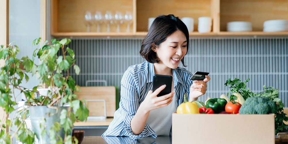 A woman holds her phone and a credit card in her kitchen with a box of produce next to her