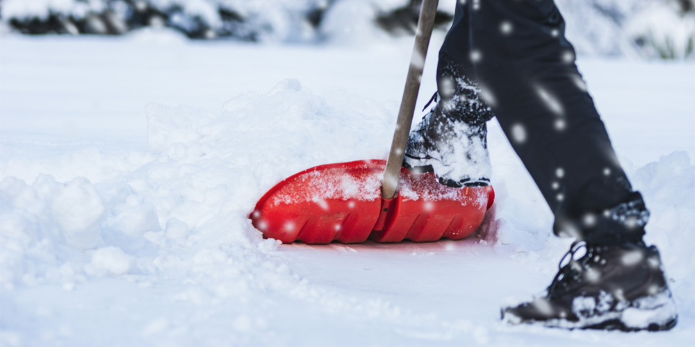 An orange shovel pushing through snow on the ground by someone wearing black pants and black boots.