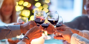 A group of people's hands holding glasses of red wine and clinking them together in a toast. Behind them is an out-of-focus christmas tree with lights.