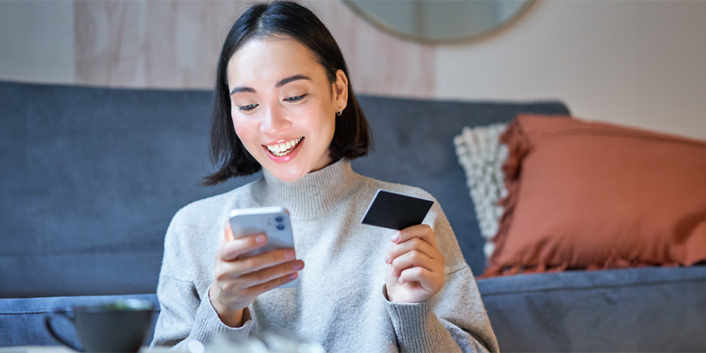 A woman sits on the floor in front of a muted blue couch wearing a grey turtleneck sweater. She is holding her smartphone in one hand and a credit card in the other.