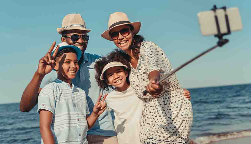 A mom, a dad and two school-aged kids in summer clothes and sun hats taking a selfie with their cellphone on a selfie stick