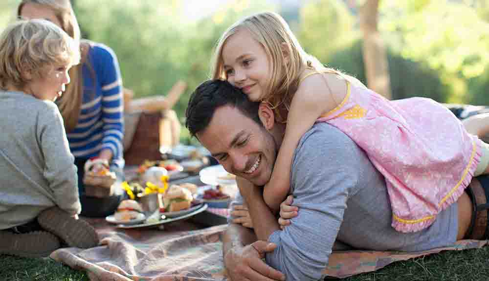 A young girl with blond hair lying on her smiling dad's back with her arms wrapped his neck. In the background is a woman sitting on a blanket with a young boy sitting in front of her. They have plates of food in front of them.
