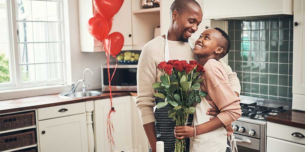 A couple are standing in a kitchen with white cabinets and a dark tile backsplash behind the range. They are each wearing aprons with their arms around each other and smiling at each other. She is holding a bouquet of red roses. There are several red heart-shaped balloons with red ribbons tied to the door knob of one of the kitchen cabinets behind them. 