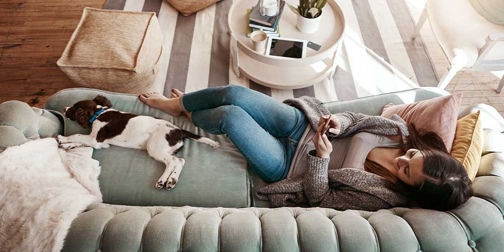 An overhead view of a woman wearing a chunky grey cardigan with a tank top and jeans lying on a light green couch. She is leaning back on two throw pillows that are pink and yellow. There is a brown and white dog lying at the other end of the couch at her feet. There is a fuzzy white throw over one end of the couch. There is a round white coffee table in front of her with a white mug of coffee or tea. There is a small potted plant on the table as well.