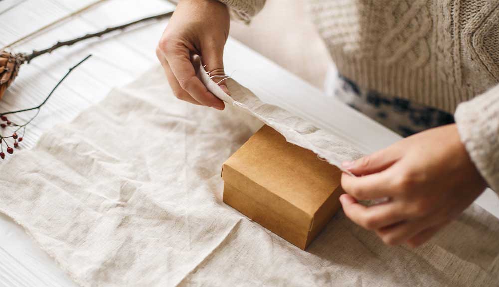 A person wearing a white knit sweater is holding a wrinkled swatch of white cloth and folding it over a brown paper box.