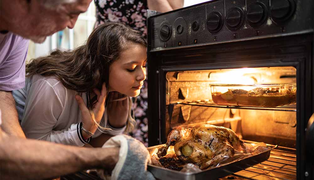 Two people are huddled over an open oven door. An older man wearing a light purple T-shirt is holding onto a silver tray with a turkey on it. A younger girl with curly brown hair is holding her hair back and leaning over the turkey.