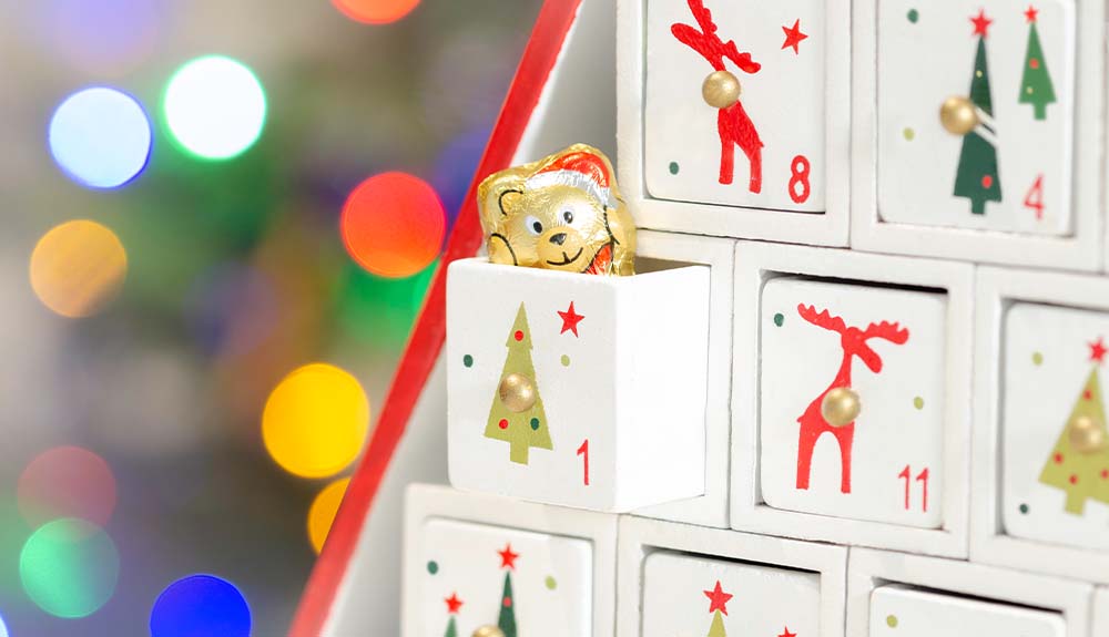 A close up view of a wooden advent calendar. Boxes 8, 4, 1 and 11 are visible. Each one has a small illustration on it, such as a Christmas tree or a reindeer. Peeking out from box one is a tinfoil wrapped chocolate bear.